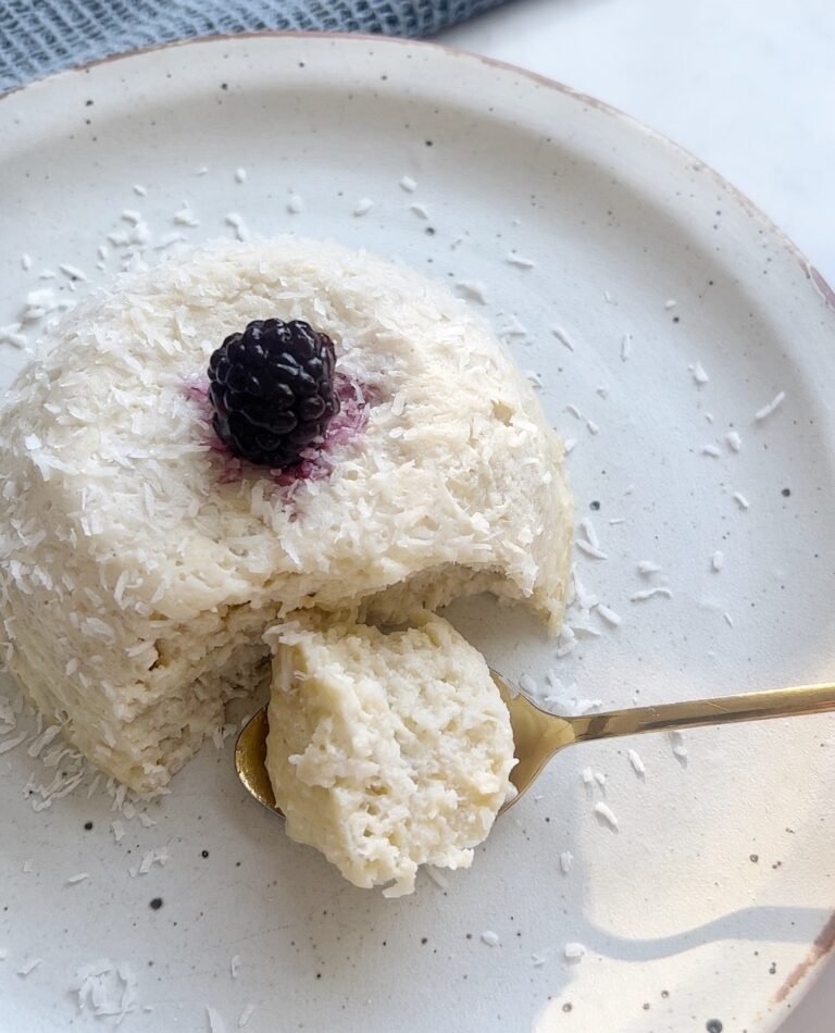 Flourless coconut cake in a ramekin topped with fresh berries and coconut flakes, served on a rustic plate.