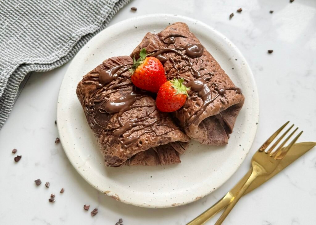 Thin chocolate crepes filled with creamy yoghurt and fresh strawberries, folded and topped with chocolate drizzle on a white plate.