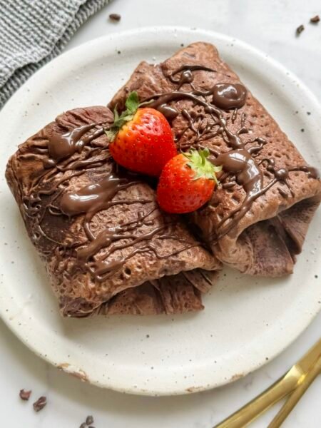 Thin chocolate crepes filled with creamy yoghurt and fresh strawberries, folded and topped with chocolate drizzle on a white plate.