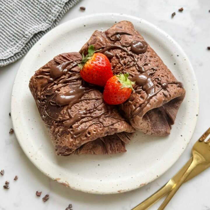 Thin chocolate crepes filled with creamy yoghurt and fresh strawberries, folded and topped with chocolate drizzle on a white plate.