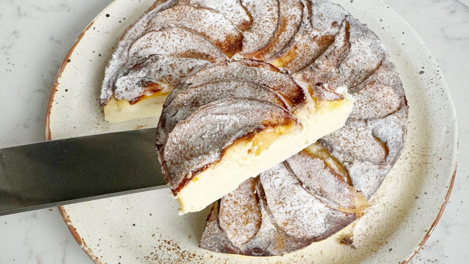 A golden-brown slice of spiced apple yoghurt cake topped with tender apple chunks and a light dusting of cinnamon. The cake looks moist and fluffy, served on a plate with a dollop of yoghurt and a sprinkle of nuts, creating a warm and wholesome presentation.