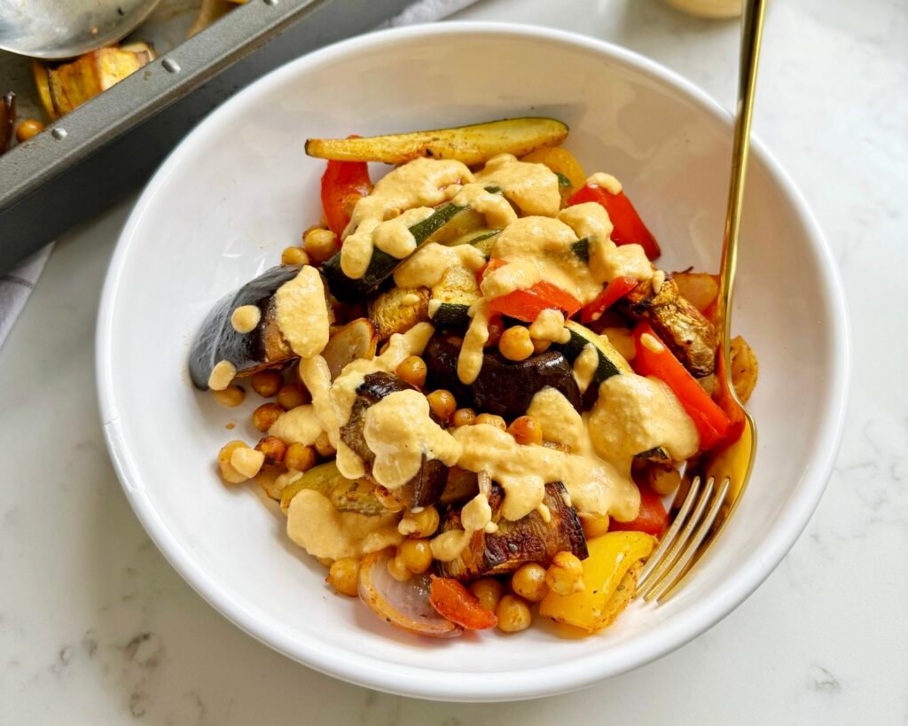 A vibrant bowl of roasted Mediterranean vegetables—aubergine, courgette, onions, peppers—tossed with chickpeas and drizzled with creamy lemon-tahini dressing.