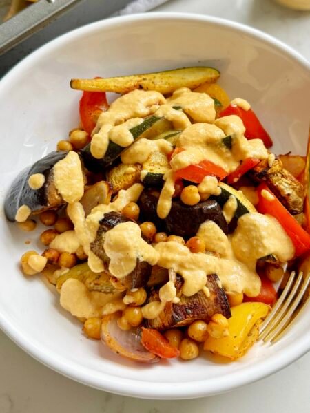 A vibrant bowl of roasted Mediterranean vegetables—aubergine, courgette, onions, peppers—tossed with chickpeas and drizzled with creamy lemon-tahini dressing.
