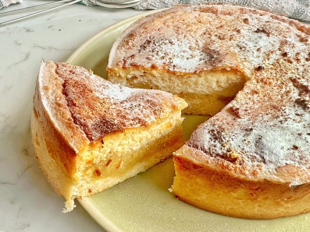 A freshly baked Apple and Almond Yoghurt Cake with a golden-brown crust, sliced to reveal a soft, moist interior with a layer of apple purée in the middle. The cake is presented on a wooden board, garnished with a sprinkle of cinnamon and slivered almonds, with a dollop of yoghurt on the side." Let me know if you'd like any adjustments!