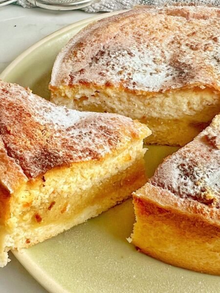 A freshly baked Apple and Almond Yoghurt Cake with a golden-brown crust, sliced to reveal a soft, moist interior with a layer of apple purée in the middle. The cake is presented on a wooden board, garnished with a sprinkle of cinnamon and slivered almonds, with a dollop of yoghurt on the side." Let me know if you'd like any adjustments!