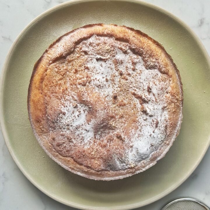 A freshly baked Apple and Almond Yoghurt Cake with a golden-brown crust, sliced to reveal a soft, moist interior with a layer of apple purée in the middle. The cake is presented on a wooden board, garnished with a sprinkle of cinnamon and slivered almonds, with a dollop of yoghurt on the side.