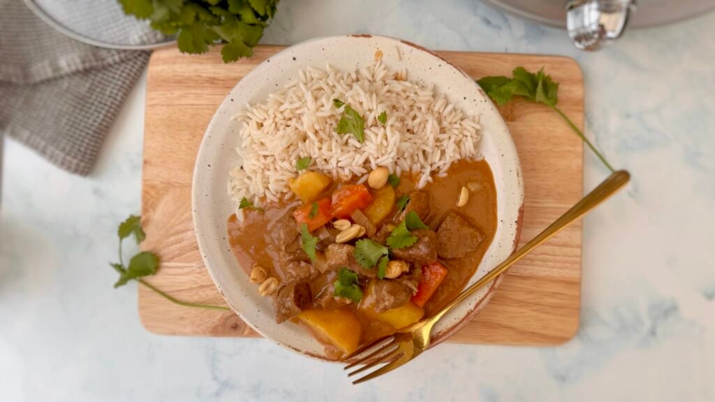 A bowl of steaming Beef Massaman Curry featuring tender beef chunks, diced potatoes, and carrots in a creamy, spiced coconut milk sauce, garnished with fresh coriander and served over jasmine rice.