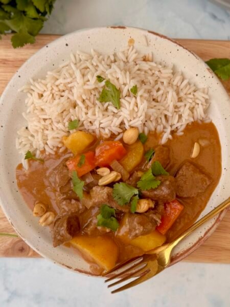 A bowl of steaming Beef Massaman Curry featuring tender beef chunks, diced potatoes, and carrots in a creamy, spiced coconut milk sauce, garnished with fresh coriander and served over jasmine rice.