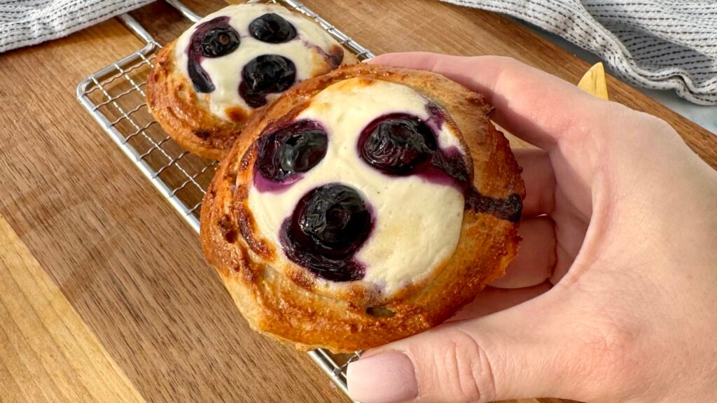 A close-up of freshly baked Blueberry Breakfast Buns with a golden-brown exterior, filled with creamy yoghurt and juicy blueberries. The buns are placed on a white plate, with a drizzle of sugar-free maple syrup and a sprinkle of fresh berries around them. The soft, fluffy texture contrasts with the vibrant blueberries, making for a delicious and healthy breakfast treat.