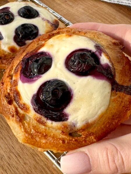 A close-up of freshly baked Blueberry Breakfast Buns with a golden-brown exterior, filled with creamy yoghurt and juicy blueberries. The buns are placed on a white plate, with a drizzle of sugar-free maple syrup and a sprinkle of fresh berries around them. The soft, fluffy texture contrasts with the vibrant blueberries, making for a delicious and healthy breakfast treat.