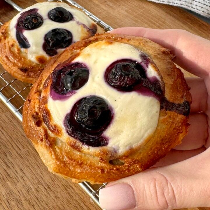 A close-up of freshly baked Blueberry Breakfast Buns with a golden-brown exterior, filled with creamy yoghurt and juicy blueberries. The buns are placed on a white plate, with a drizzle of sugar-free maple syrup and a sprinkle of fresh berries around them. The soft, fluffy texture contrasts with the vibrant blueberries, making for a delicious and healthy breakfast treat.