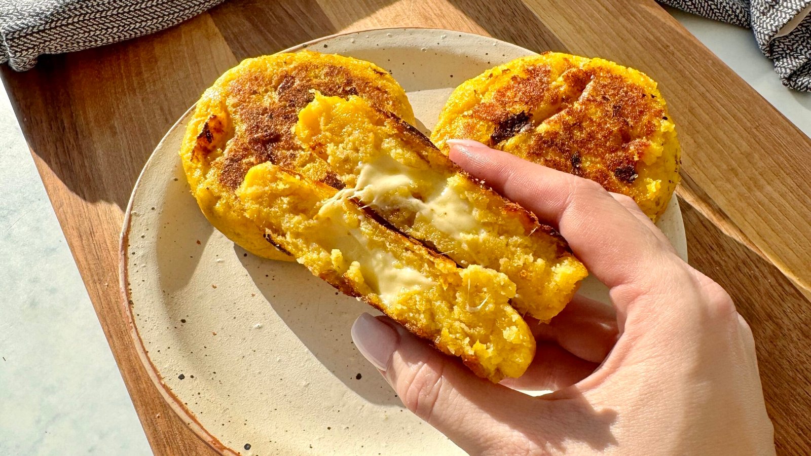 Crispy, golden-brown cheese-stuffed plantain arepas with a soft, sweet interior and gooey melted cheese filling, served warm on a plate with avocado slices and a side of dipping sauce.
