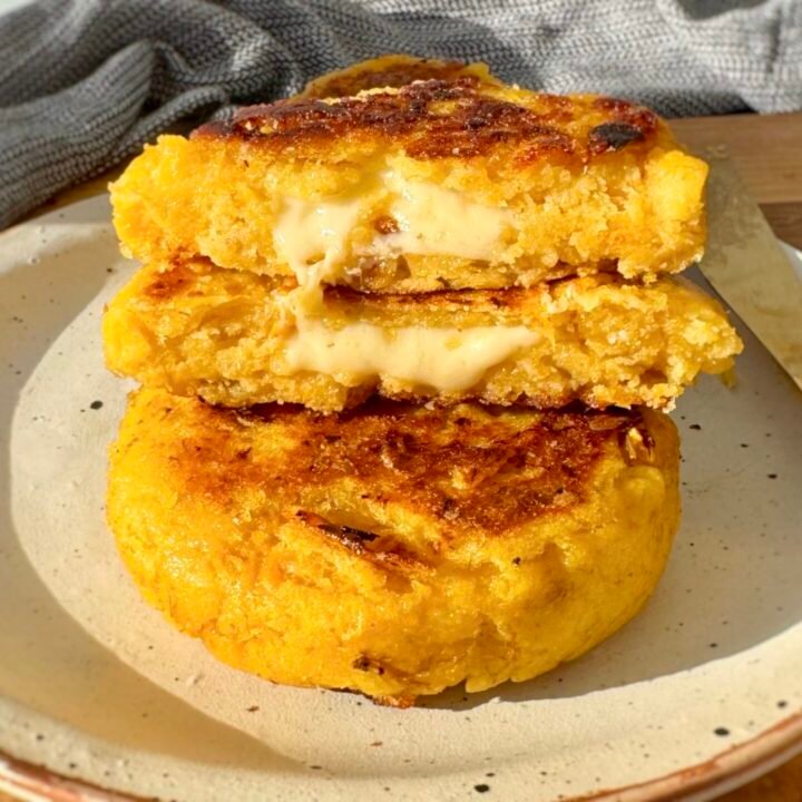 Crispy, golden-brown cheese-stuffed plantain arepas with a soft, sweet interior and gooey melted cheese filling, served warm on a plate with avocado slices and a side of dipping sauce.