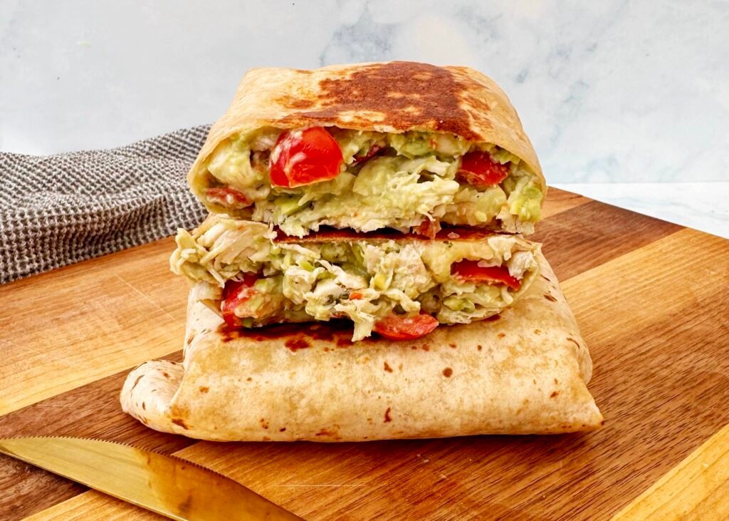 A golden, toasted Chicken Avocado Burrito cut in half, revealing tender shredded chicken, creamy avocado, melted cheese, and fresh diced tomatoes inside a whole wheat tortilla. Served on a plate with a side of Greek yogurt and garnished with fresh herbs.