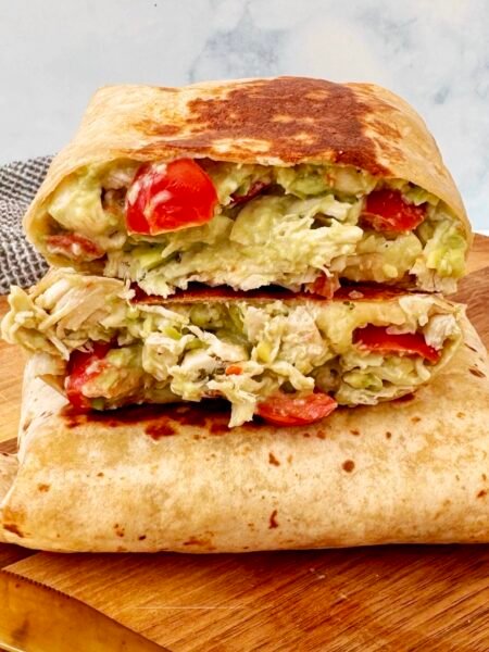 A golden, toasted Chicken Avocado Burrito cut in half, revealing tender shredded chicken, creamy avocado, melted cheese, and fresh diced tomatoes inside a whole wheat tortilla. Served on a plate with a side of Greek yogurt and garnished with fresh herbs.