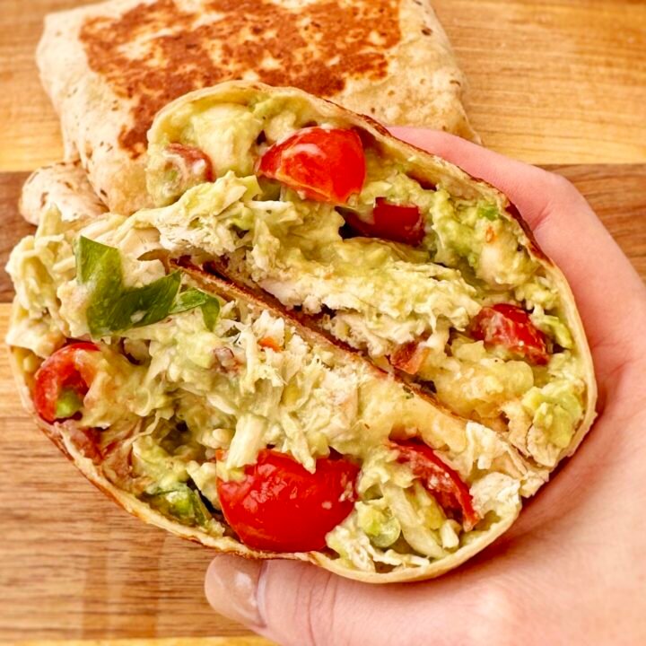 A golden, toasted Chicken & Avocado Burrito cut in half, revealing tender shredded chicken, creamy avocado, melted cheese, and fresh diced tomatoes inside a whole wheat tortilla. Served on a plate with a side of Greek yogurt and garnished with fresh herbs.
