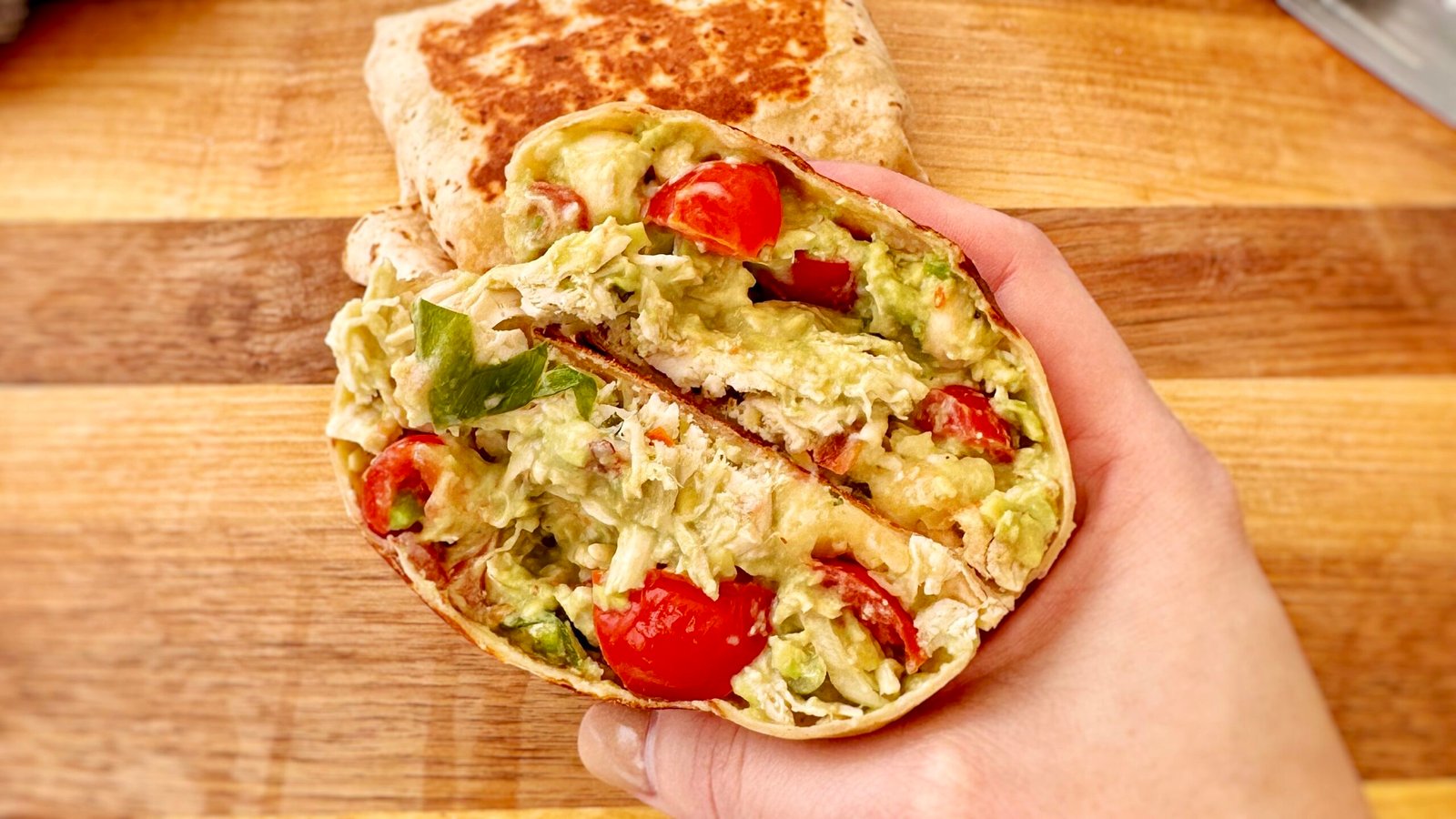 A golden, toasted Chicken & Avocado Burrito cut in half, revealing tender shredded chicken, creamy avocado, melted cheese, and fresh diced tomatoes inside a whole wheat tortilla. Served on a plate with a side of Greek yogurt and garnished with fresh herbs.