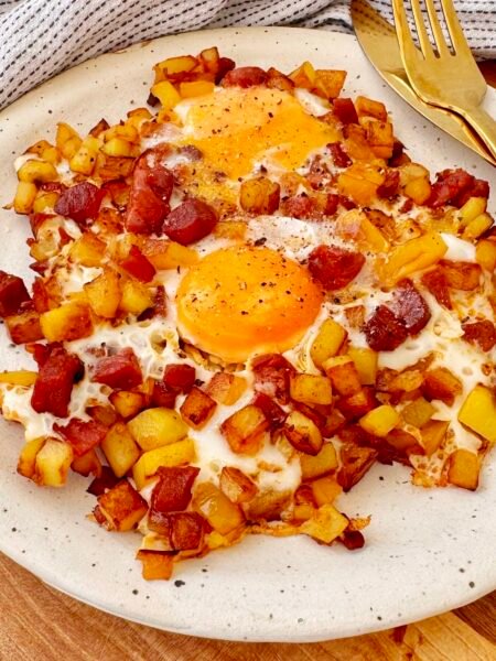 A hearty and flavorful Chorizo Egg Hash served in a skillet. Crispy golden-brown potatoes, smoky chorizo, and perfectly cooked eggs with runny yolks are garnished with paprika and fresh herbs. The dish is vibrant with yellow bell peppers and caramelized onions, making it a delicious and satisfying one-pan meal.