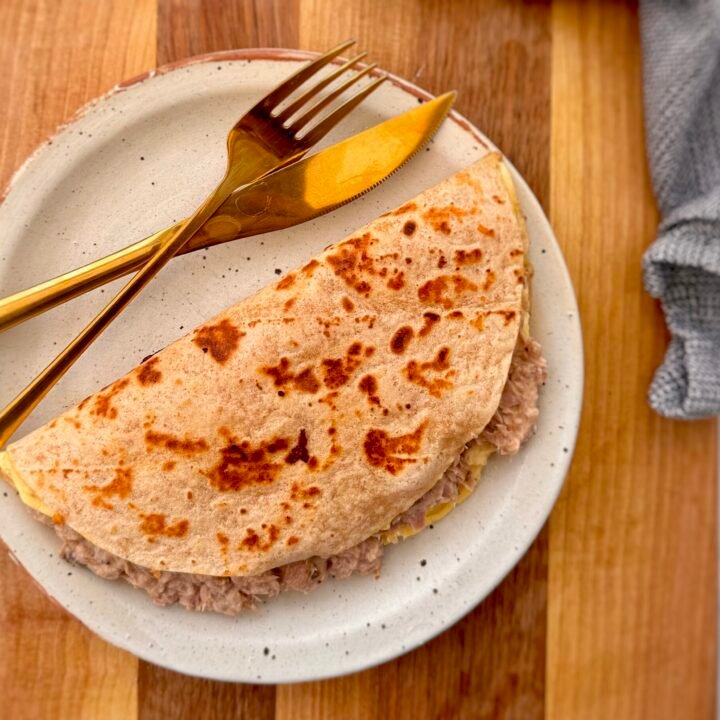 A golden-brown tortilla omelette folded in half, filled with creamy cottage cheese and a savory tuna mixture. The omelette sits on a plate, garnished with fresh herbs, with a side of salad and a drizzle of hot sauce for extra flavor.