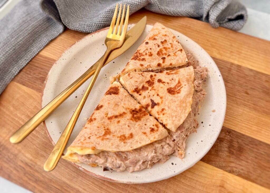 A golden-brown tortilla omelette folded in half, filled with creamy cottage cheese and a savory tuna mixture. The omelette sits on a plate, garnished with fresh herbs, with a side of salad and a drizzle of hot sauce for extra flavor.