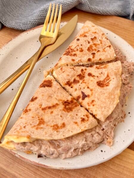 A golden-brown tortilla omelette folded in half, filled with creamy cottage cheese and a savory tuna mixture. The omelette sits on a plate, garnished with fresh herbs, with a side of salad and a drizzle of hot sauce for extra flavor.