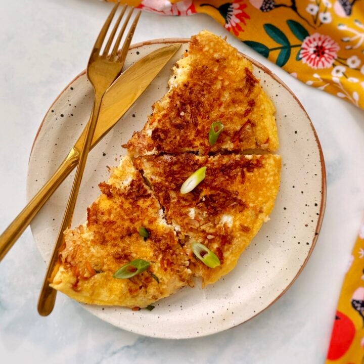 : A golden, crispy rice and carrot omelette folded in half, revealing a fluffy egg interior with bits of grated carrot and crispy rice. Garnished with chopped spring onions and served on a plate with a side of fresh salad.