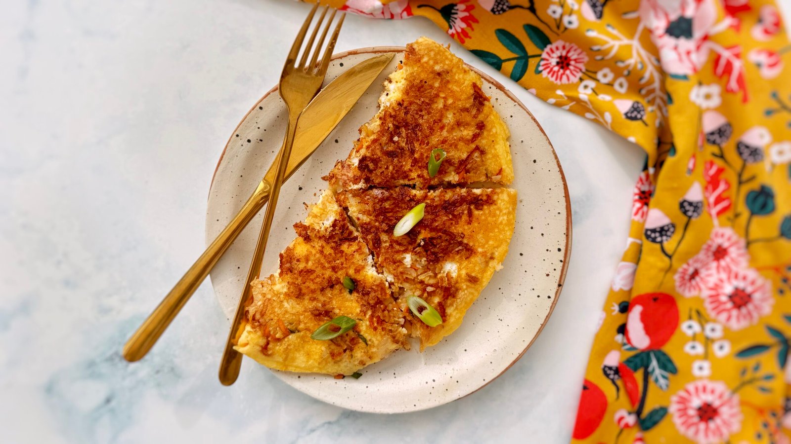 : A golden, crispy rice and carrot omelette folded in half, revealing a fluffy egg interior with bits of grated carrot and crispy rice. Garnished with chopped spring onions and served on a plate with a side of fresh salad.