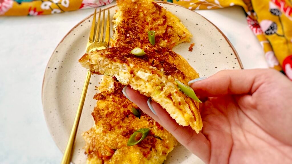 : A golden, crispy rice and carrot omelette folded in half, revealing a fluffy egg interior with bits of grated carrot and crispy rice. Garnished with chopped spring onions and served on a plate with a side of fresh salad.