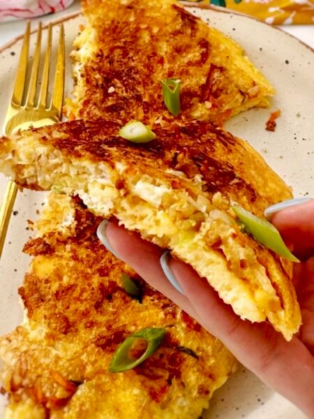 : A golden, crispy rice and carrot omelette folded in half, revealing a fluffy egg interior with bits of grated carrot and crispy rice. Garnished with chopped spring onions and served on a plate with a side of fresh salad.