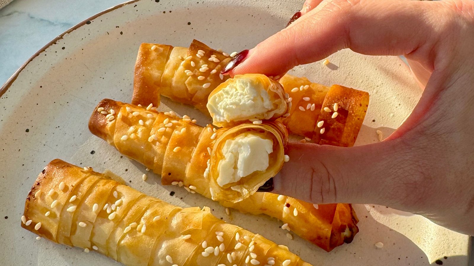 "Golden-brown filo pastries with a crispy, flaky texture, drizzled with glistening hot honey and sprinkled with sesame seeds. The pastries are neatly arranged on a serving plate, with one cut open to reveal creamy feta cheese inside. A small dish of extra hot honey and a few red chilli flakes are placed nearby, hinting at the sweet and spicy flavours of the dish