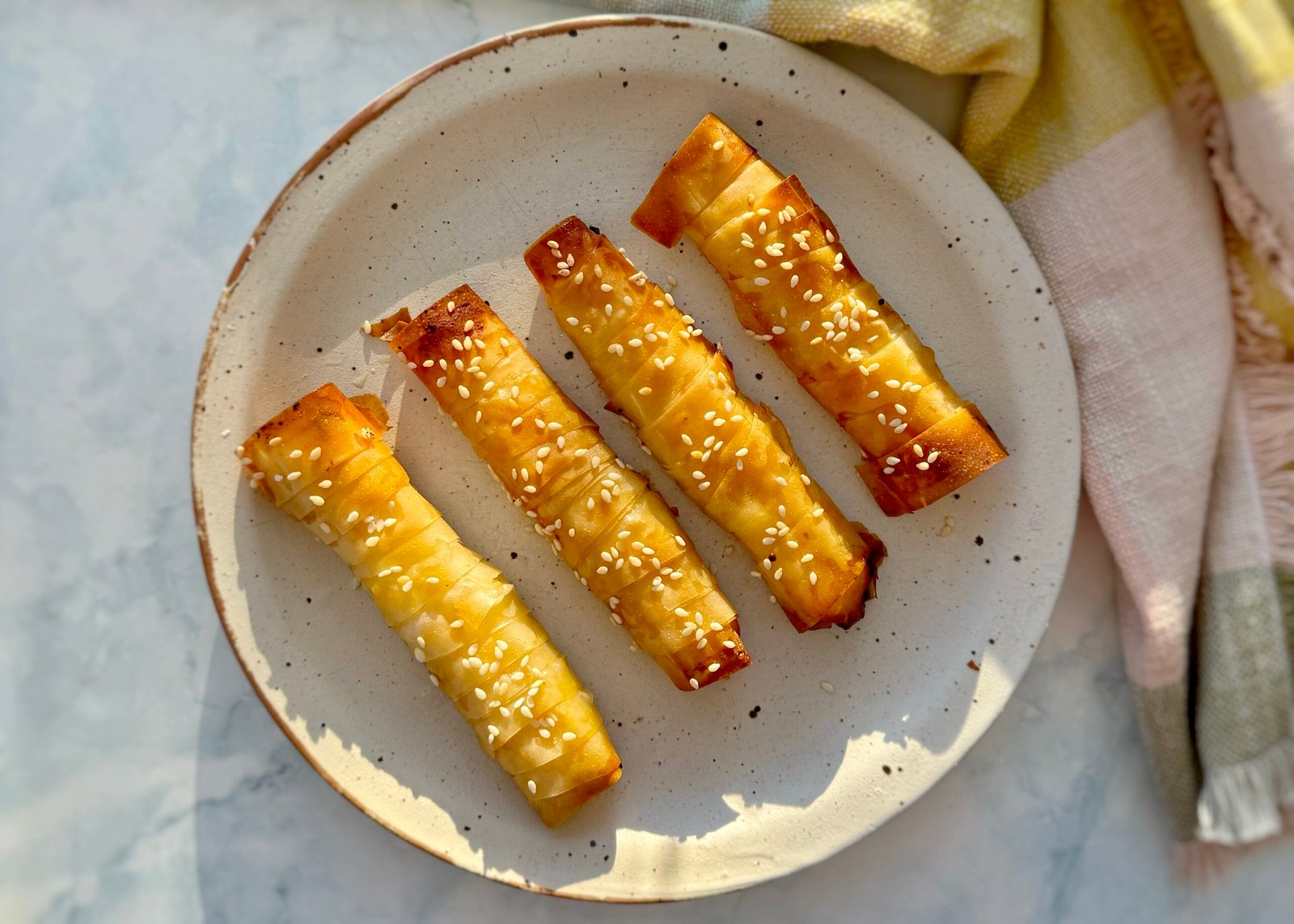 "Golden-brown filo pastries with a crispy, flaky texture, drizzled with glistening hot honey and sprinkled with sesame seeds. The pastries are neatly arranged on a serving plate, with one cut open to reveal creamy feta cheese inside. A small dish of extra hot honey and a few red chilli flakes are placed nearby, hinting at the sweet and spicy flavours of the dish