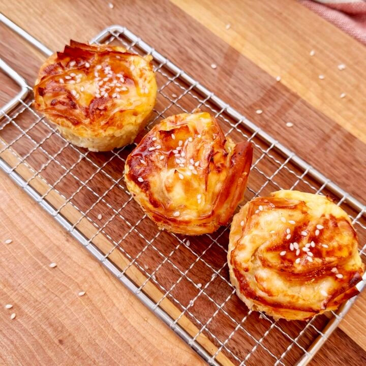 A golden, crispy filo pastry nest filled with creamy feta custard, drizzled with glistening hot honey. The delicate, crinkled layers of pastry create a light, airy texture, topped with sesame seeds for an extra crunch. Served in a muffin tin or on a rustic wooden board, these mini crinkle pies are an irresistible blend of sweet and savoury, perfect for appetizers or brunch.