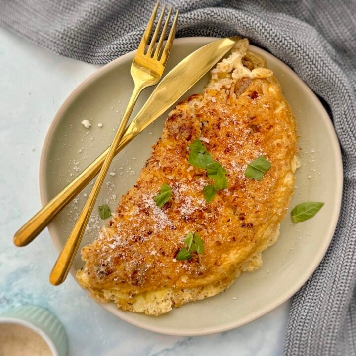 A golden-brown omelette folded over a flavorful filling of pesto-infused rice and vegetarian sausage, garnished with freshly grated Parmesan cheese. Served on a white plate with a side of fresh greens.