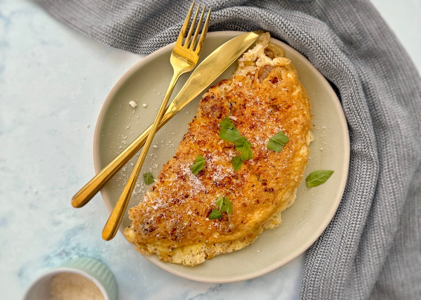 A golden-brown omelette folded over a flavorful filling of pesto-infused rice and vegetarian sausage, garnished with freshly grated Parmesan cheese. Served on a white plate with a side of fresh greens.