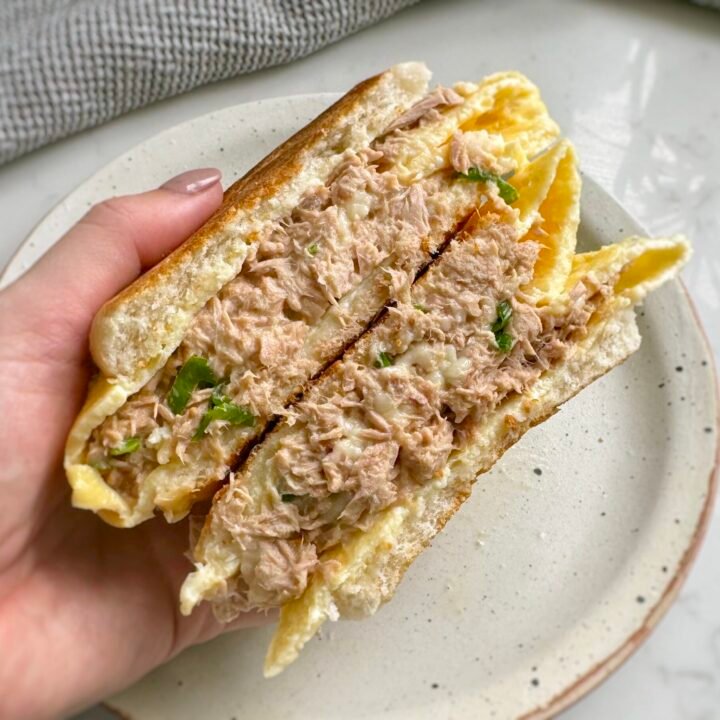 A golden toasted Tuna Egg Bap with melted cheese, creamy tuna filling, and fluffy eggs, served warm on a plate. The bread roll is crispy on the outside and soft inside, with layers of rich, savory flavors.