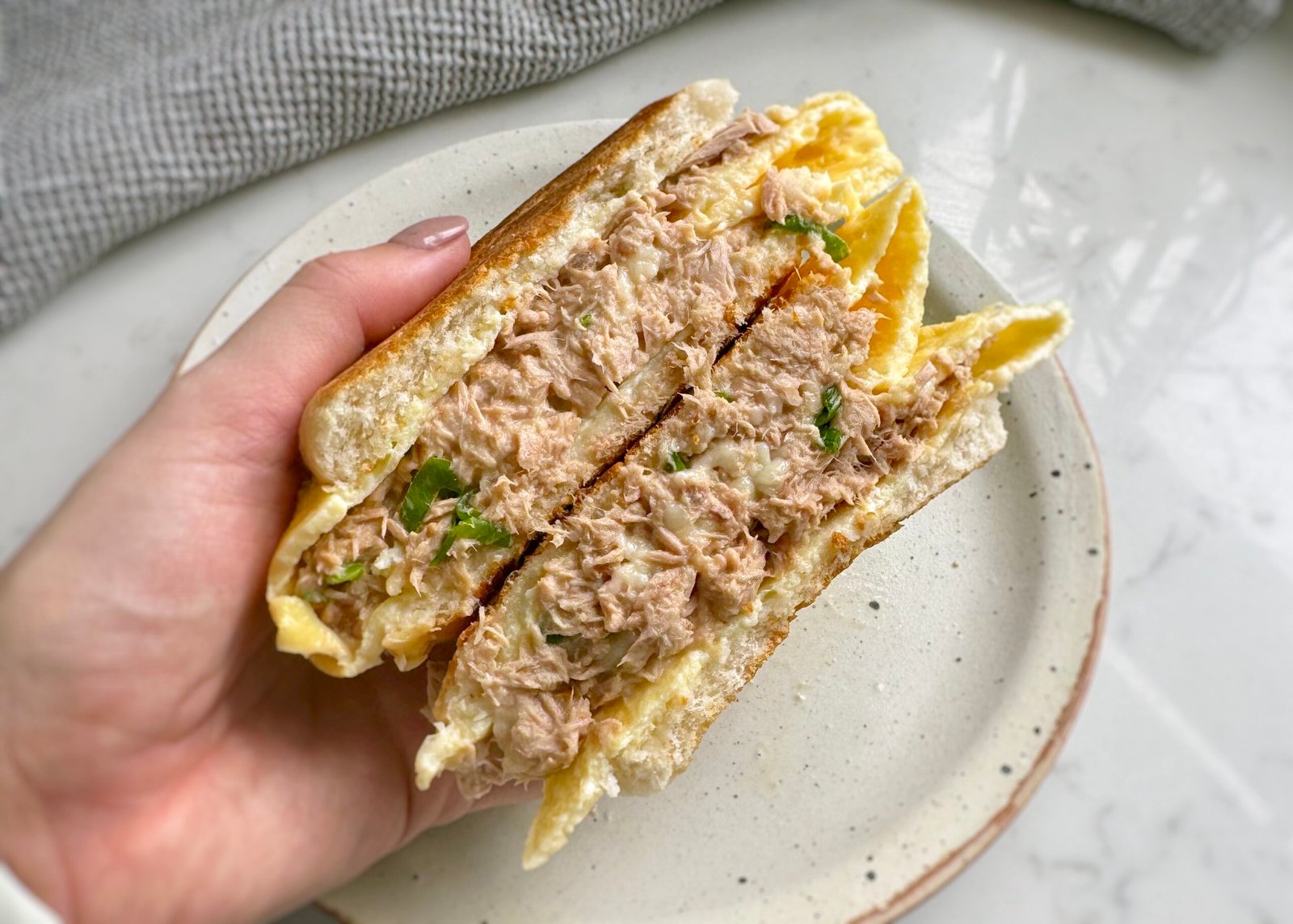 A golden toasted Tuna Egg Bap with melted cheese, creamy tuna filling, and fluffy eggs, served warm on a plate. The bread roll is crispy on the outside and soft inside, with layers of rich, savory flavors.