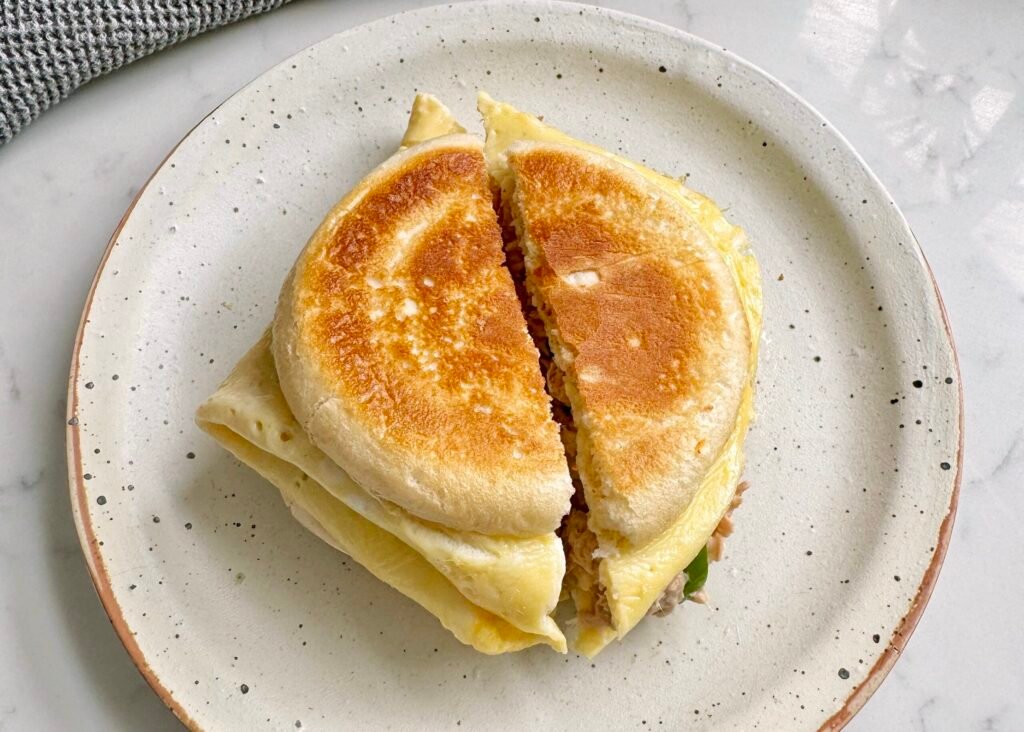 A golden toasted Tuna Egg Bap with melted cheese, creamy tuna filling, and fluffy eggs, served warm on a plate. The bread roll is crispy on the outside and soft inside, with layers of rich, savory flavors.