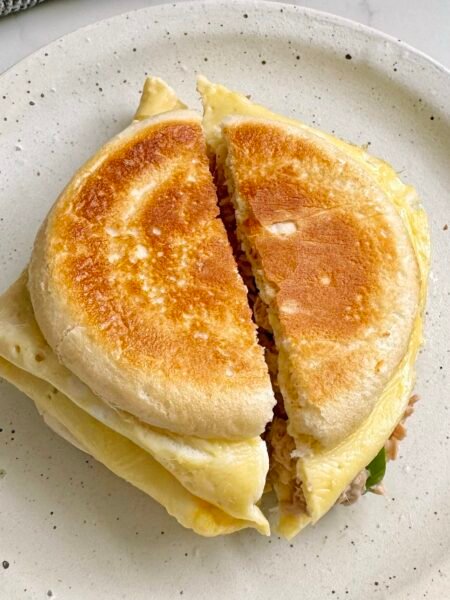 A golden toasted Tuna Egg Bap with melted cheese, creamy tuna filling, and fluffy eggs, served warm on a plate. The bread roll is crispy on the outside and soft inside, with layers of rich, savory flavors.