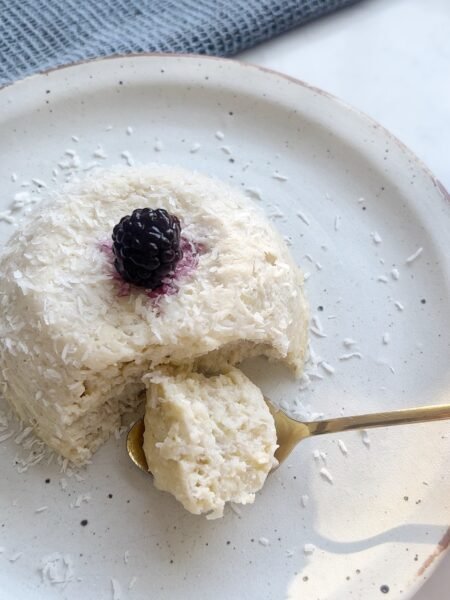 Flourless coconut cake in a ramekin topped with fresh berries and coconut flakes, served on a rustic plate.
