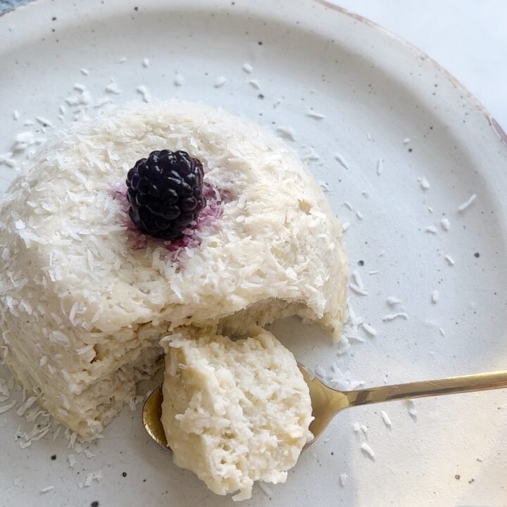 Flourless coconut cake in a ramekin topped with fresh berries and coconut flakes, served on a rustic plate.