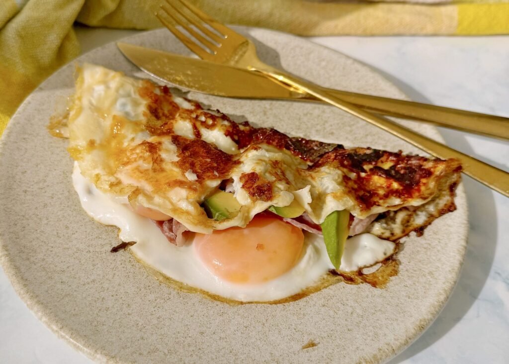 A folded cheesy egg dish topped with crispy bacon and creamy avocado slices, drizzled with hot honey, served on a rustic white plate.