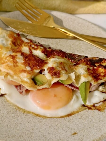 A folded cheesy egg dish topped with crispy bacon and creamy avocado slices, drizzled with hot honey, served on a rustic white plate.