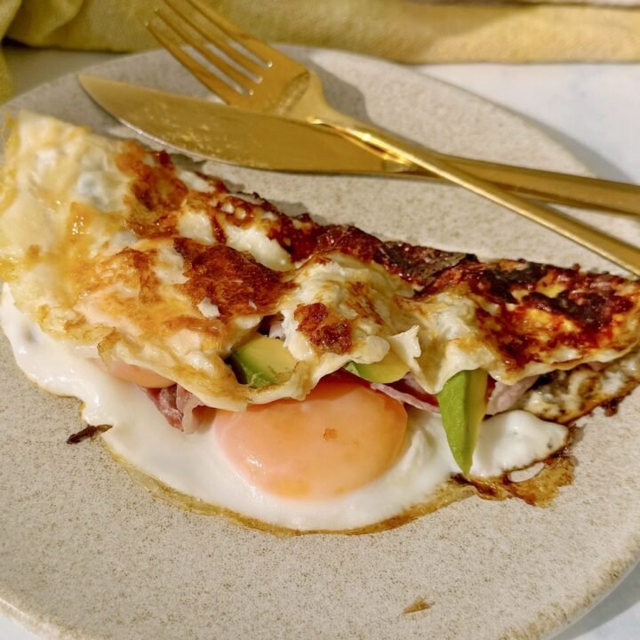 A folded cheesy egg dish topped with crispy bacon and creamy avocado slices, drizzled with hot honey, served on a rustic white plate.