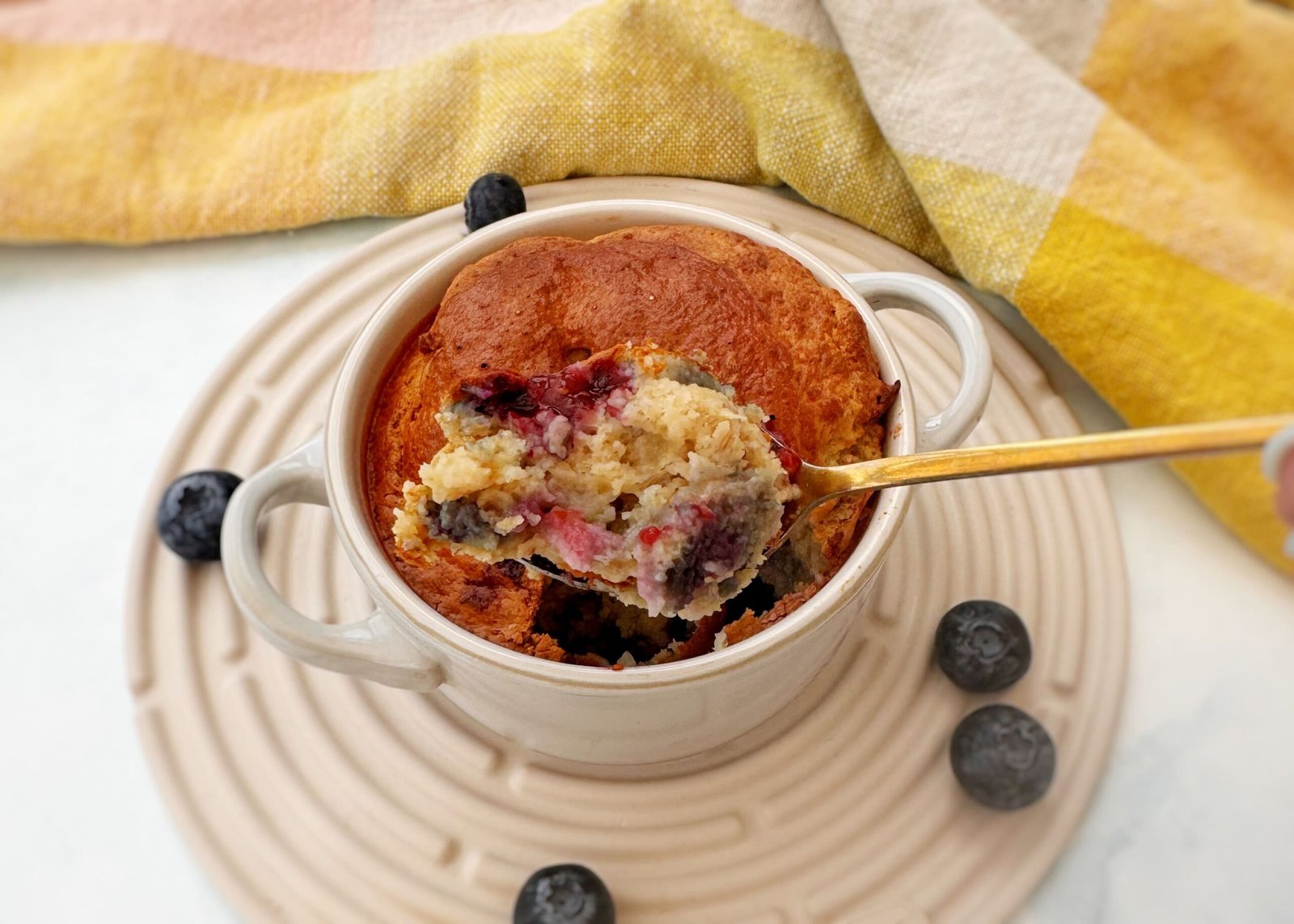 A golden-baked oatmeal dish studded with blueberries, served warm in a small white ramekin with a spoon on the side