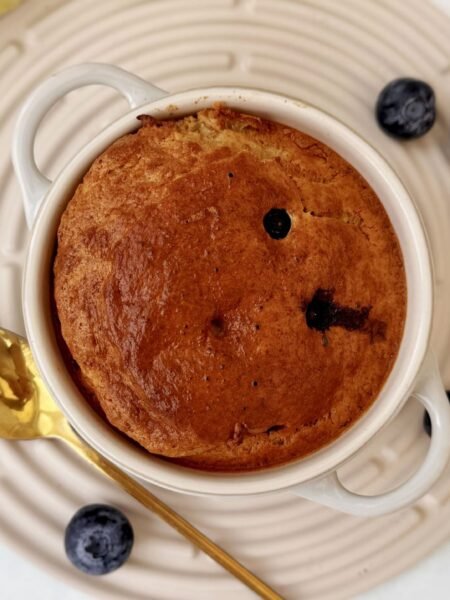 A golden-baked oatmeal dish studded with blueberries, served warm in a small white ramekin with a spoon on the side