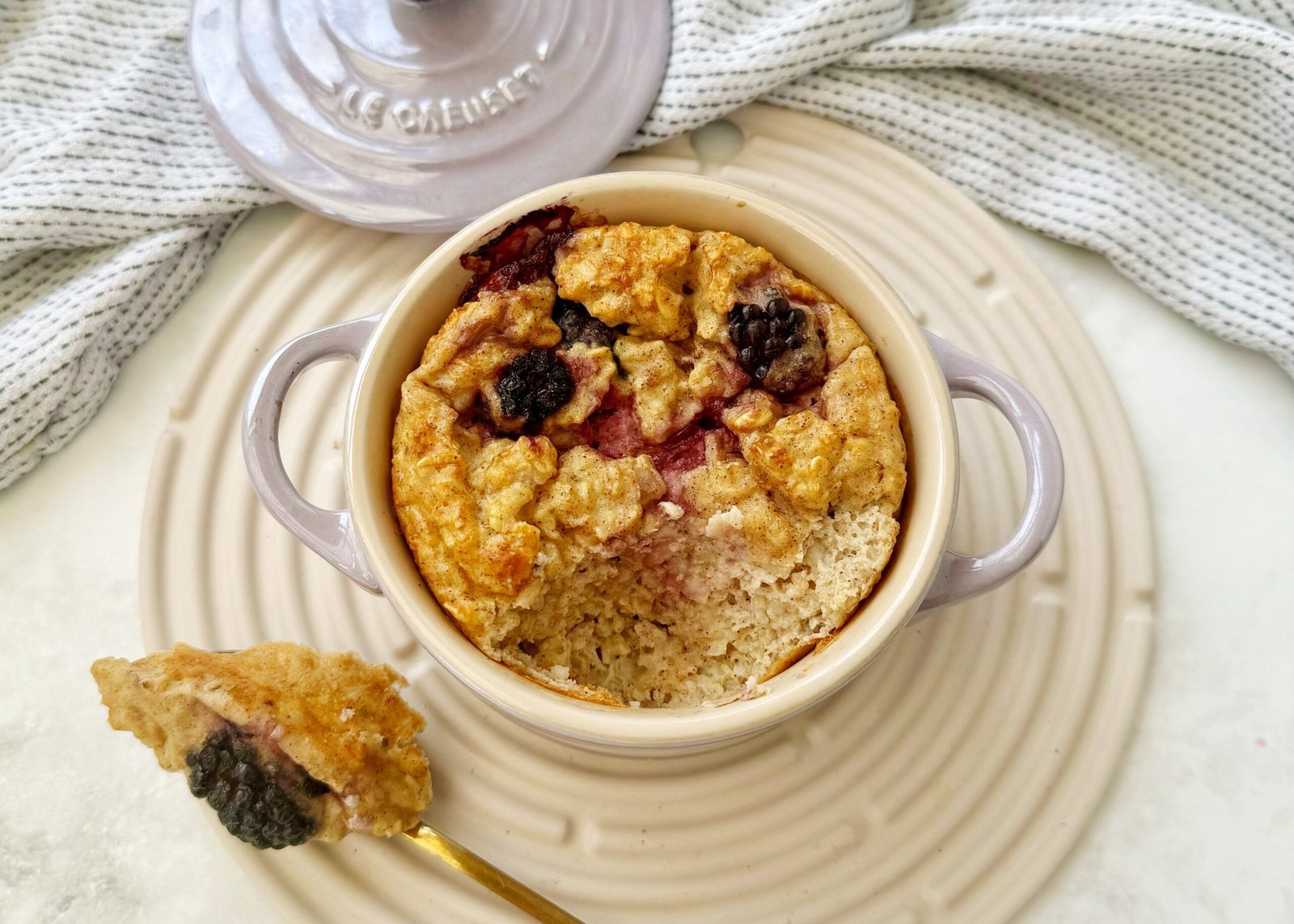 A single-serving dish of golden baked oats topped with juicy blackberries, served warm in a white ramekin with a spoon on the side.