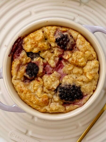 A single-serving dish of golden baked oats topped with juicy blackberries, served warm in a white ramekin with a spoon on the side.