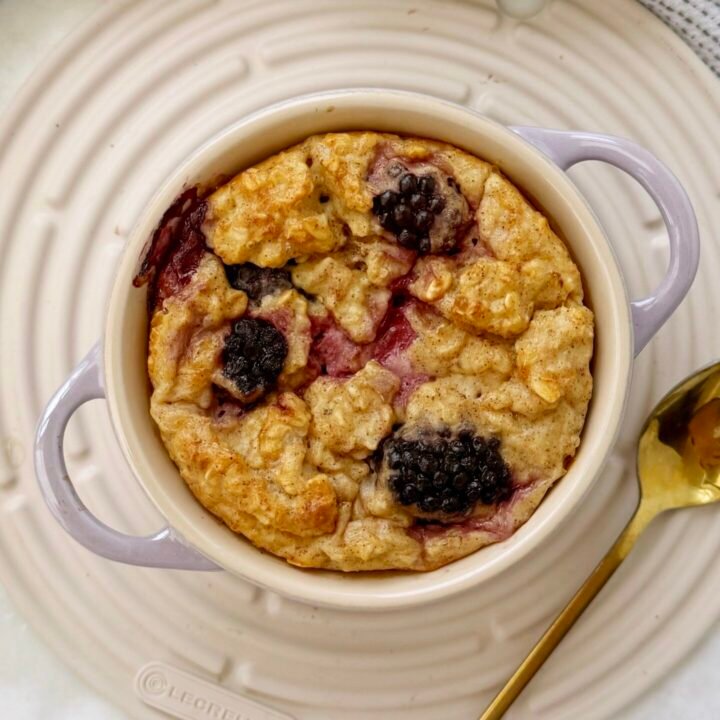 A single-serving dish of golden baked oats topped with juicy blackberries, served warm in a white ramekin with a spoon on the side.