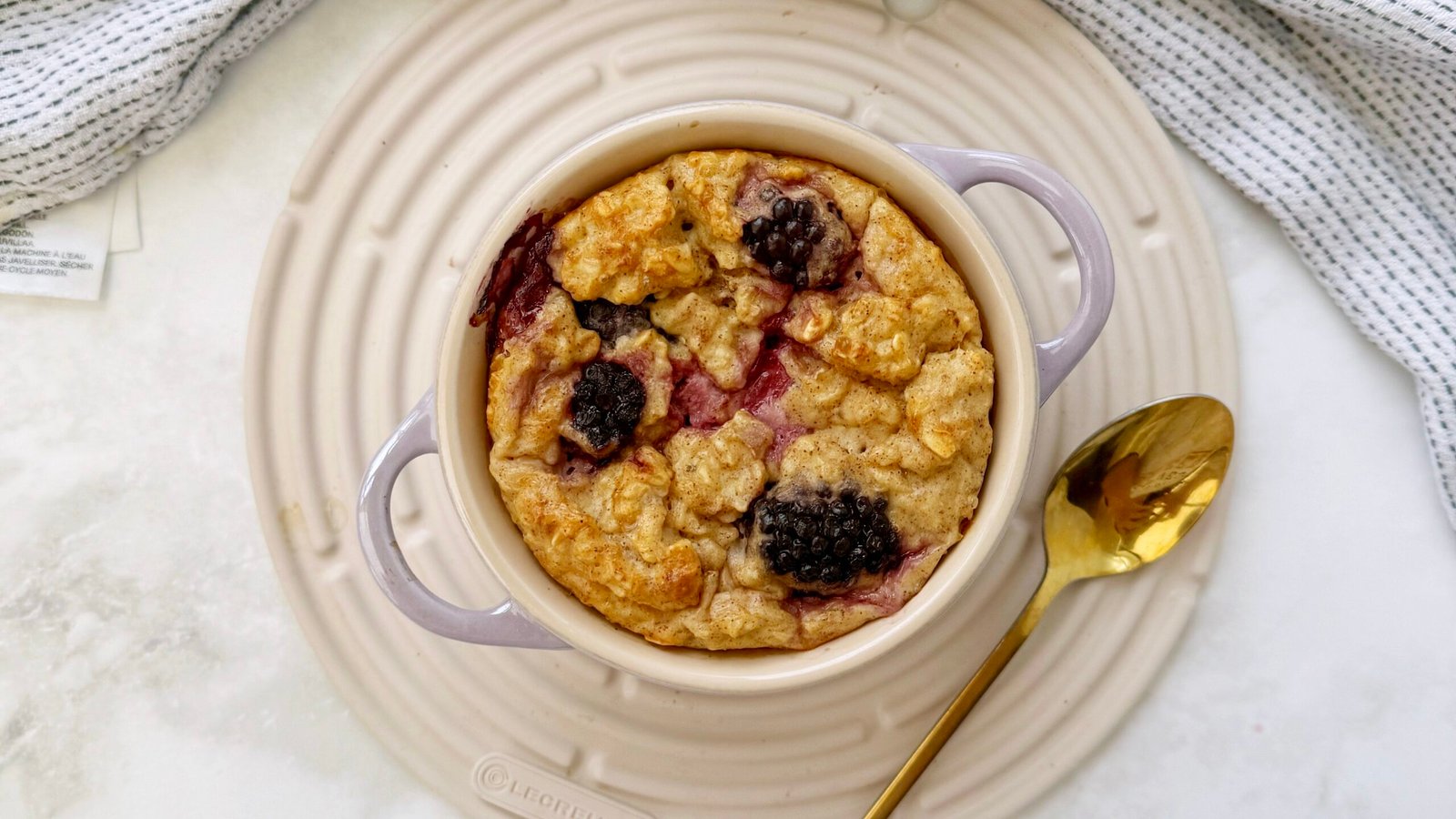 A single-serving dish of golden baked oats topped with juicy blackberries, served warm in a white ramekin with a spoon on the side.