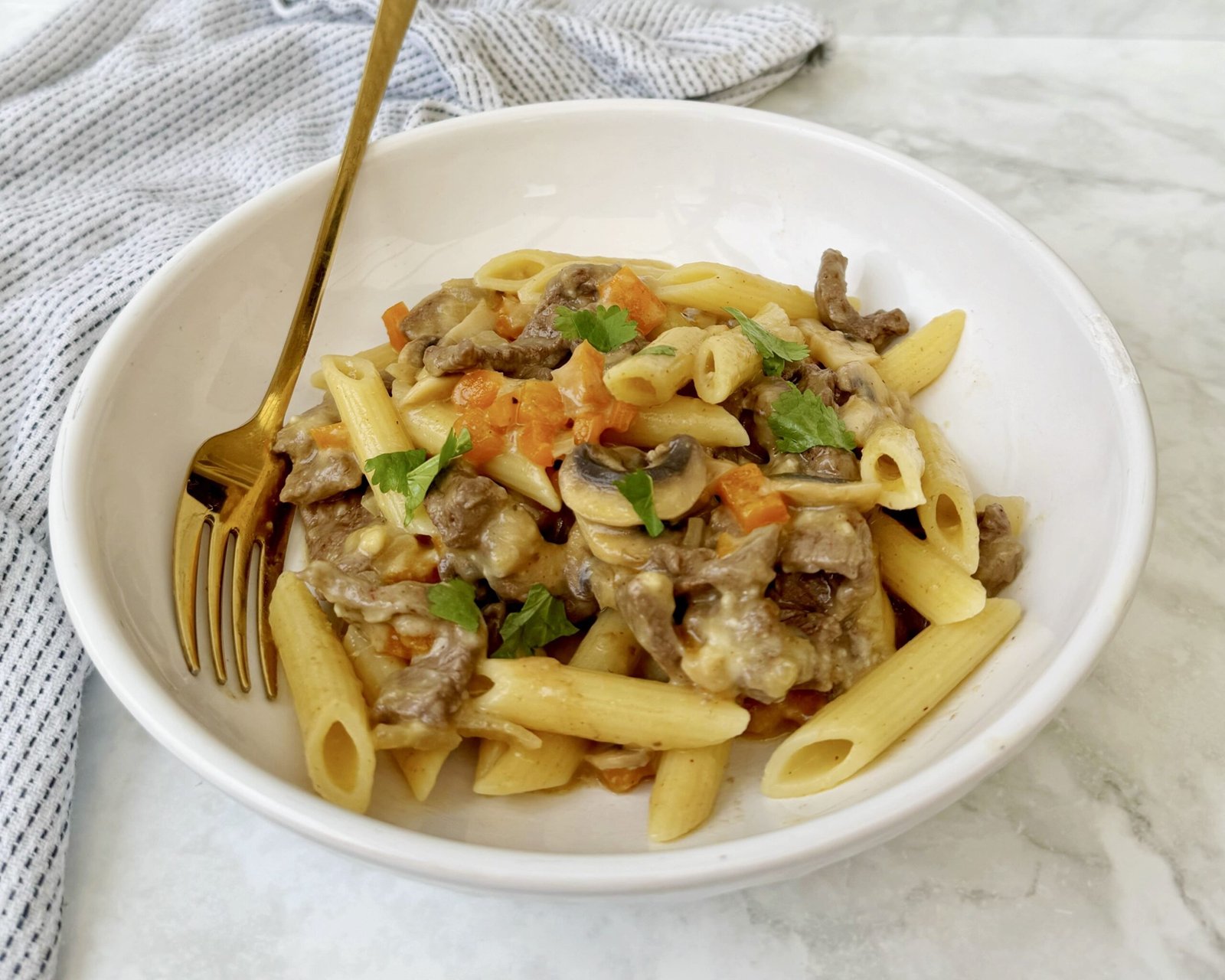 A hearty plate of cheesesteak pasta featuring thin slices of seared beef, colorful sautéed peppers, mushrooms, and onions, all tossed with whole wheat pasta in a creamy, cheesy sauce.